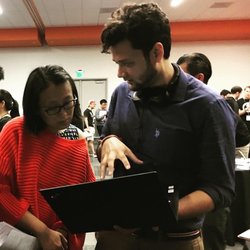 Archit holds a laptop open and points at the screen while an attendee in a red sweater reads along, in a busy conference hall.